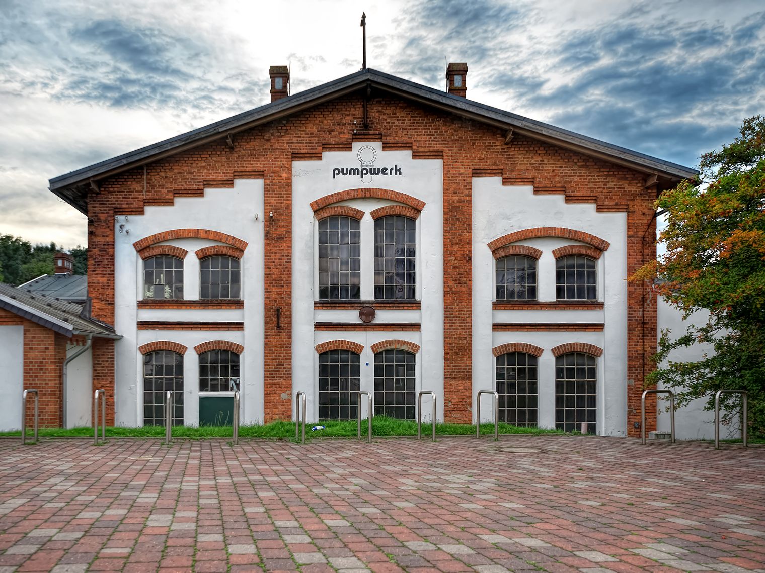 Frontansicht des Kulturzentrums Pumpwerk mit roter Ziegelfassade, weißen Wandflächen und gepflastertem Vorplatz.