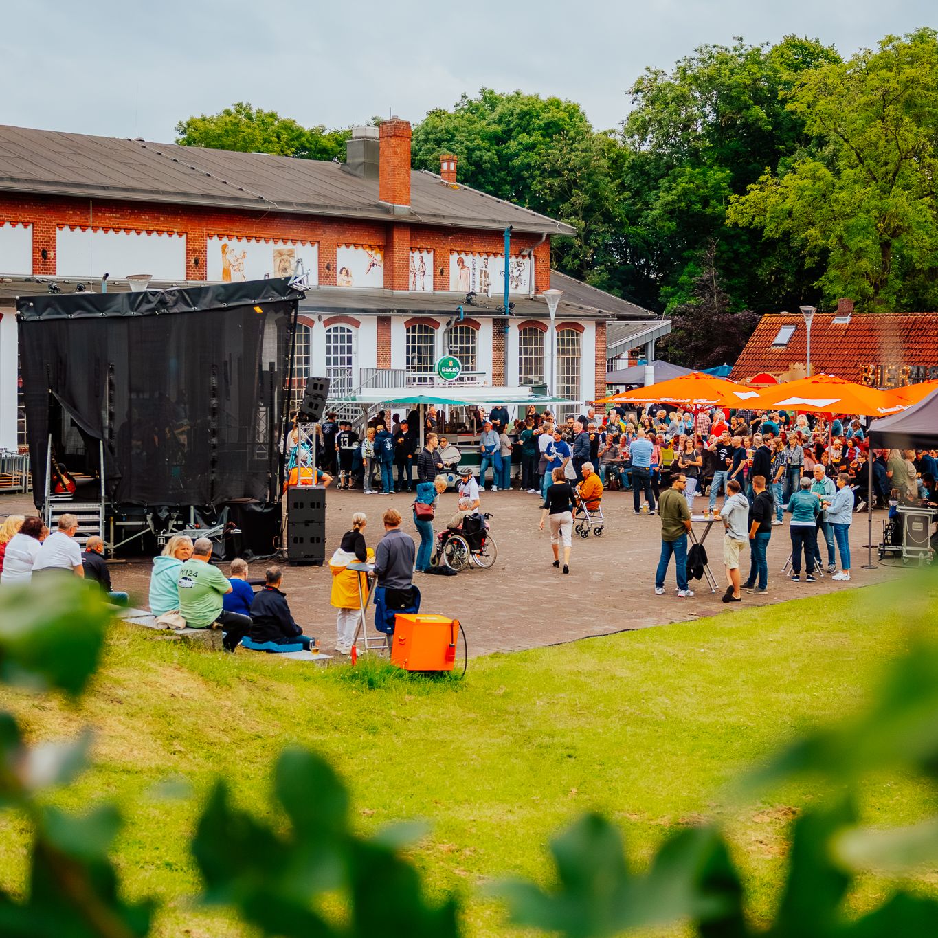 Viele Menschen bei einer Outdoor-Veranstaltung vor einem Backsteingebäude mit Bühne und Sonnenschirmen.