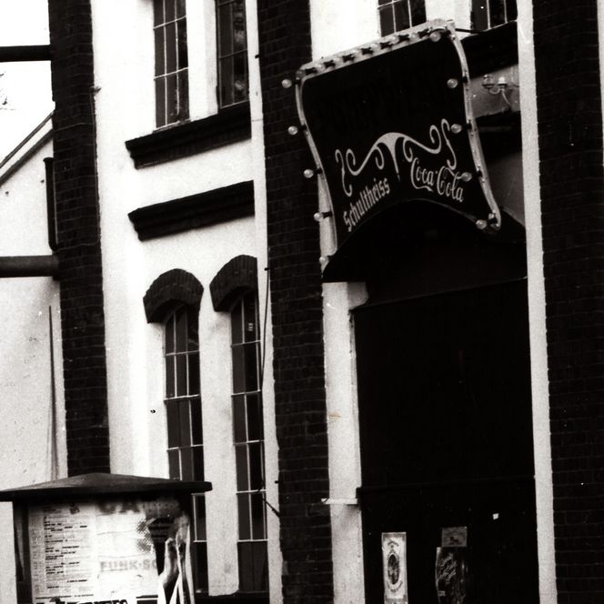 Eingang des Pumpwerks Wilhelmshaven in den 1990er Jahren, mit Werbeschild über der Tür und Litfaßsäule mit Plakaten im Vordergrund.