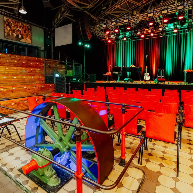 Blick in den Pumpwerk-Saal mit Bühne, roter Bestuhlung und historischem Schwungrad im Vordergrund.