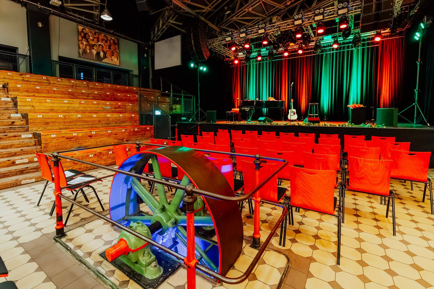 Blick in den Pumpwerk-Saal mit Bühne, roter Bestuhlung und historischem Schwungrad im Vordergrund.