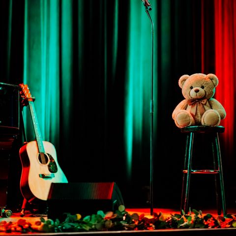 Bühne mit Mikrofonständer, Akustikgitarre und Teddybär auf einem Hocker vor farbig beleuchtetem Vorhang.