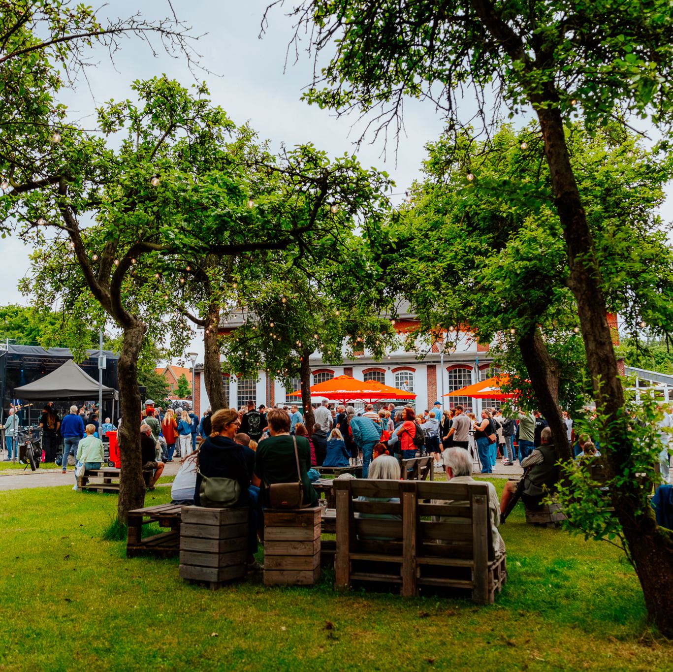 Viele Menschen bei einer Open-Air-Veranstaltung am Pumpwerk mit Bühne, Bäumen und Sitzplätzen auf der Wiese.