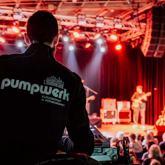 Techniker am Mischpult mit Blick auf eine rot beleuchtete Bühne und Band im Pumpwerk